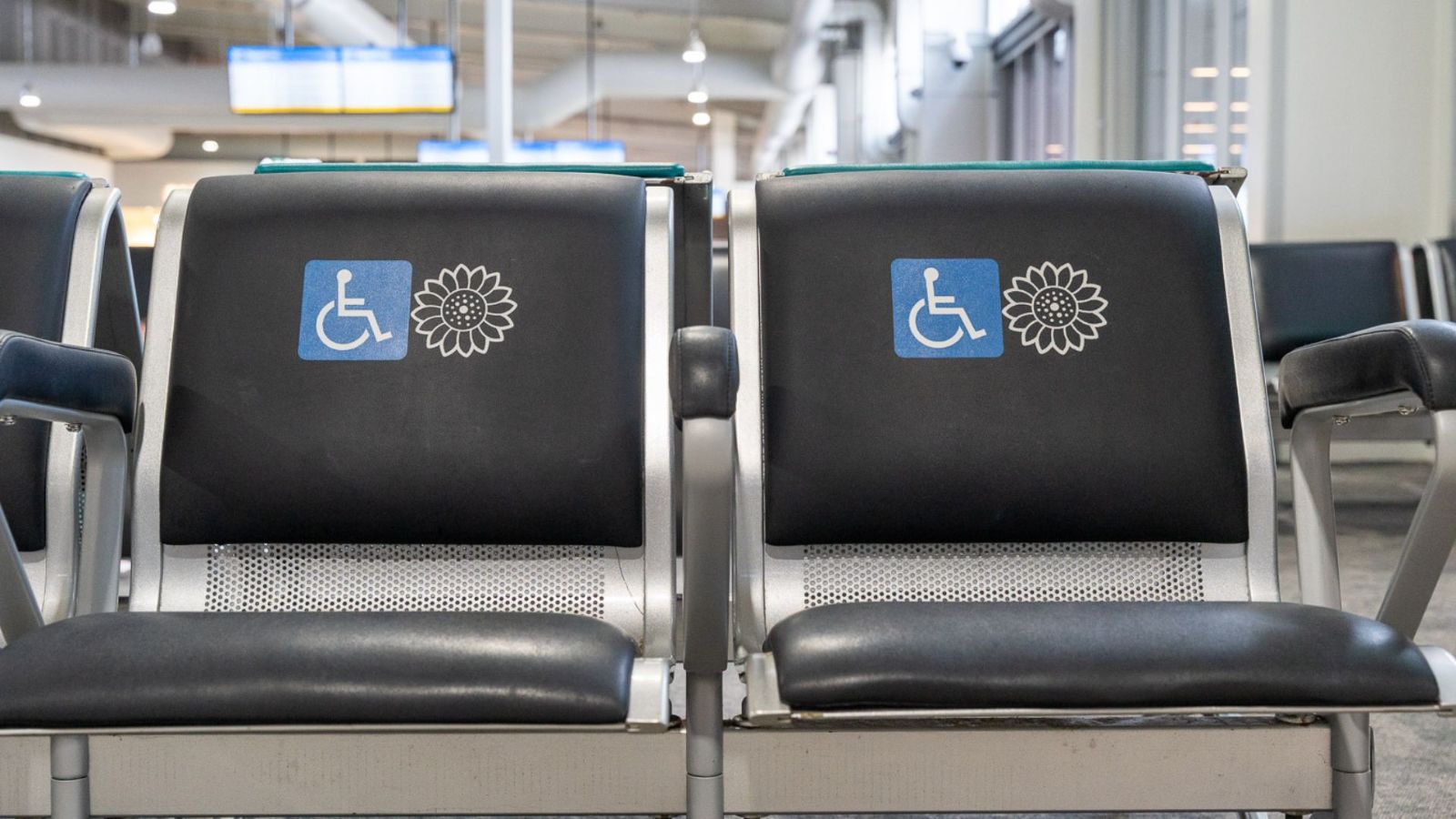 Two black airport terminal seats marked with blue accessibility symbols and Sunflower lanyard icons, indicating priority seating for travellers with disabilities or hidden disabilities.