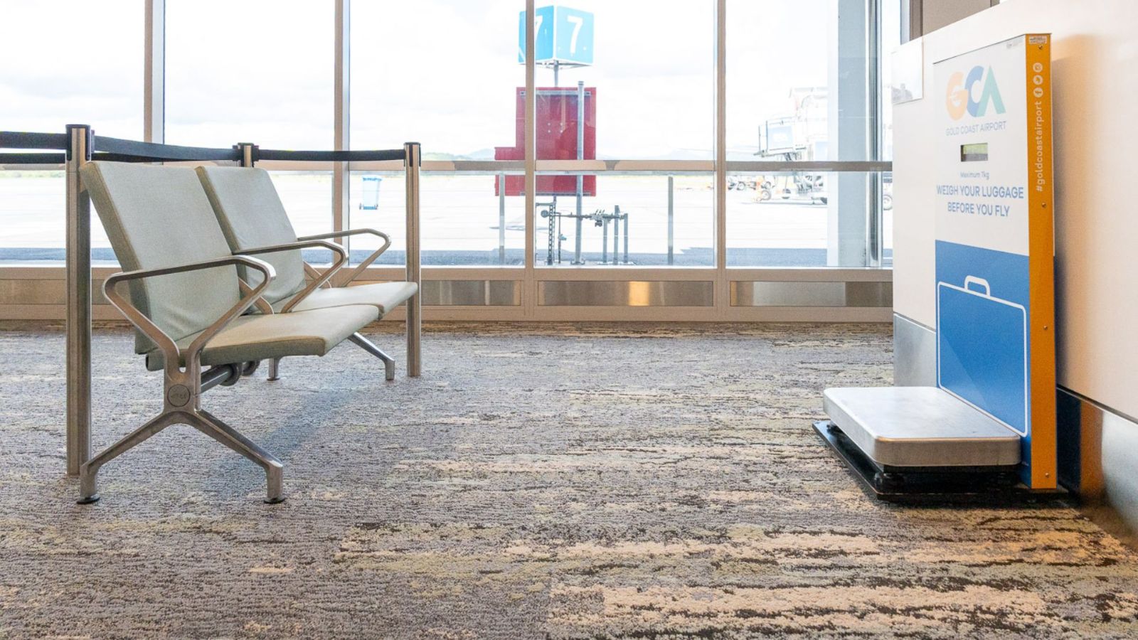Airport gate area featuring a quiet seating zone next to an accessibility-friendly baggage scale marked with the Gold Coast Airport logo. Large windows offer views of the tarmac and boarding gates.