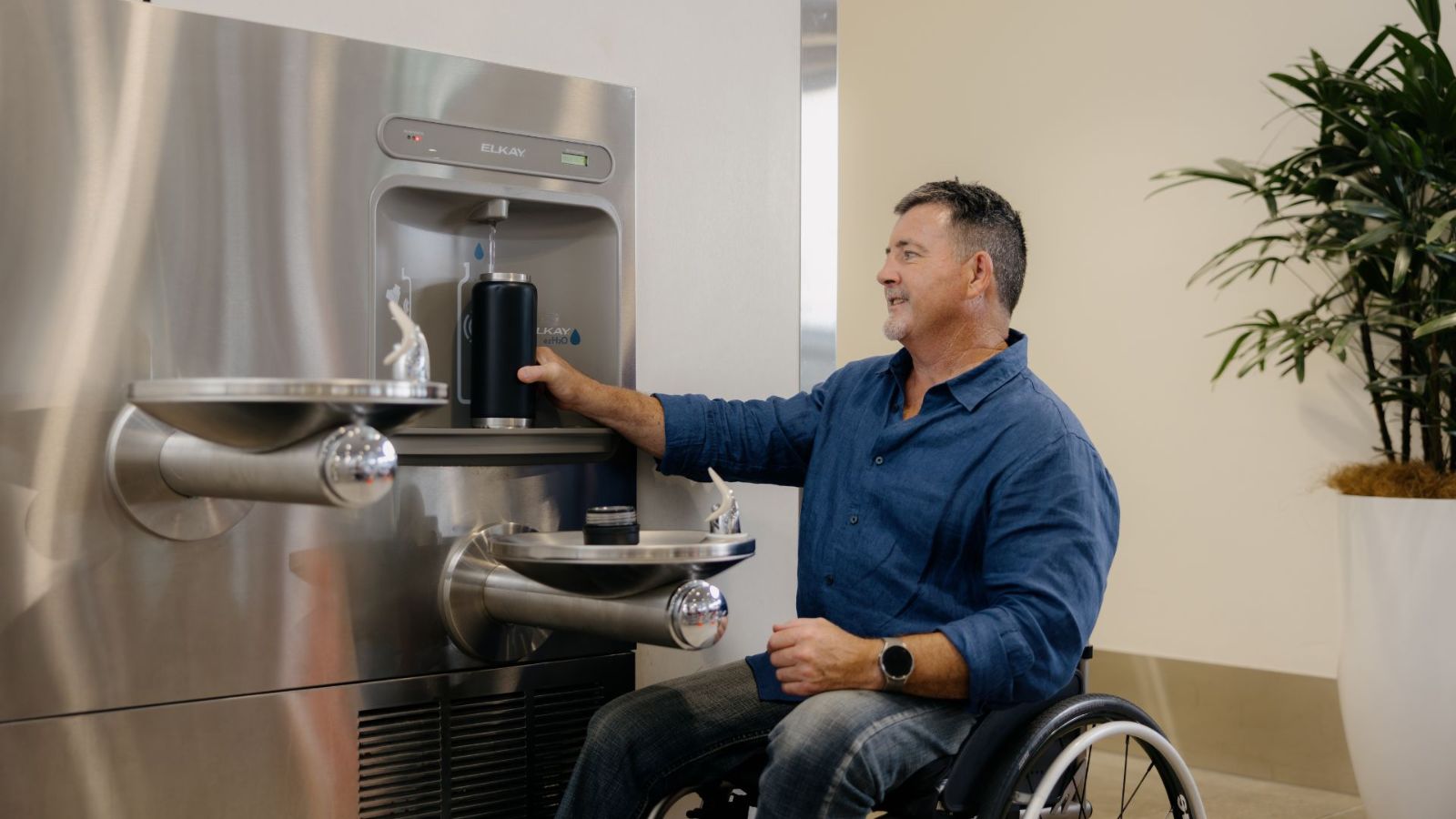 A man in a wheelchair fills his reusable water bottle at an airport hydration station, which includes dual-height drinking fountains and a bottle refill outlet. A potted plant and natural lighting enhance the welcoming space.
