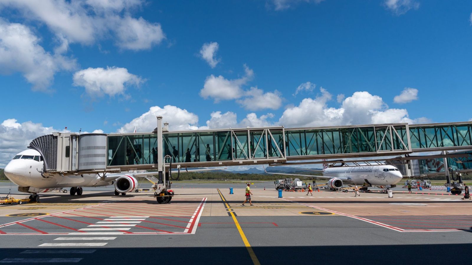 Two aircraft are parked on the tarmac at Gold Coast Airport, connected by an elevated glass jet bridge. Ground crew and vehicles operate nearby under a clear blue sky with scattered clouds.