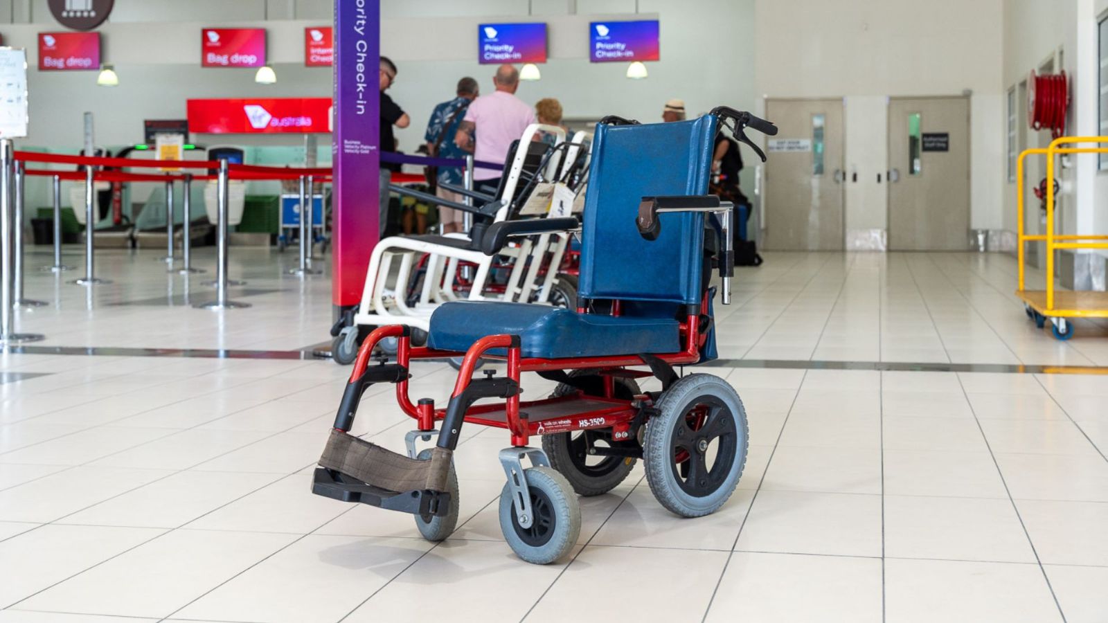 An airport wheelchair positioned on a tiled floor near check-in counters. The red and blue mobility chair is ready for passenger assistance, with people queuing and airline signage visible in the background.