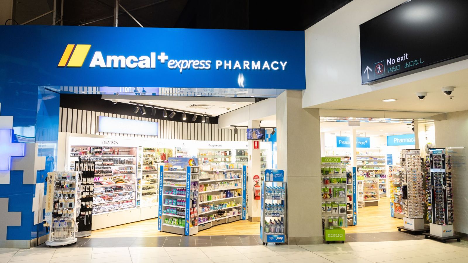 The exterior of Amcal+ Express Pharmacy at an airport, featuring a bright blue and white storefront with shelves stocked with health, wellness, and travel essentials. Display stands are positioned near the entrance.