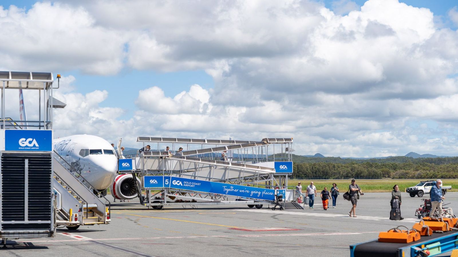 Passengers board a commercial aircraft at Gold Coast Airport via outdoor ramps. Blue GCA-branded signage is visible, with scenic mountain views in the background.