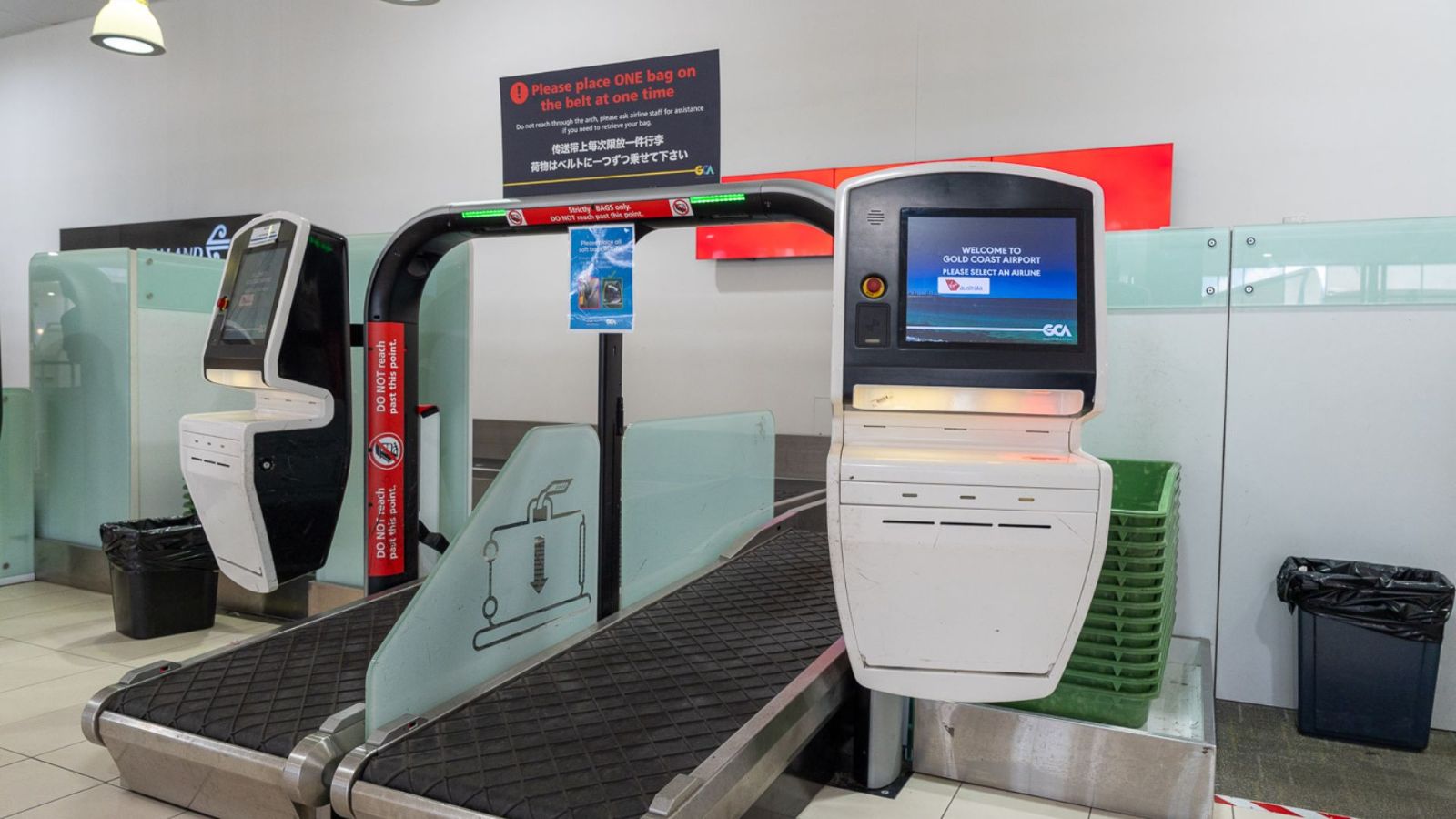 Airport self-service bag drop stations featuring conveyor belts, touchscreen kiosks, and instructional signage. The setup includes accessible design elements and privacy screens, with bins positioned nearby for convenience.