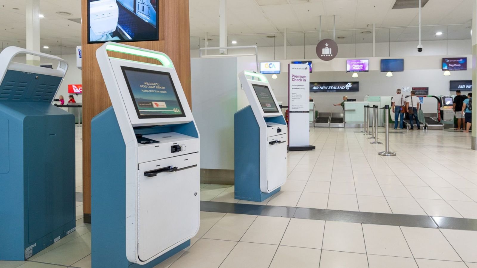A row of self-service check-in kiosks at an airport, positioned on a tiled floor near airline counters. The touchscreen machines are accessible in height and layout, with overhead screens and passengers in the background.