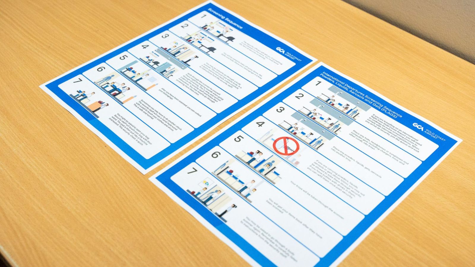 Two printed visual guides with step-by-step instructions displayed on a wooden table. The guides use blue borders and illustrations to explain airport security or screening procedures in an easy-to-follow format.