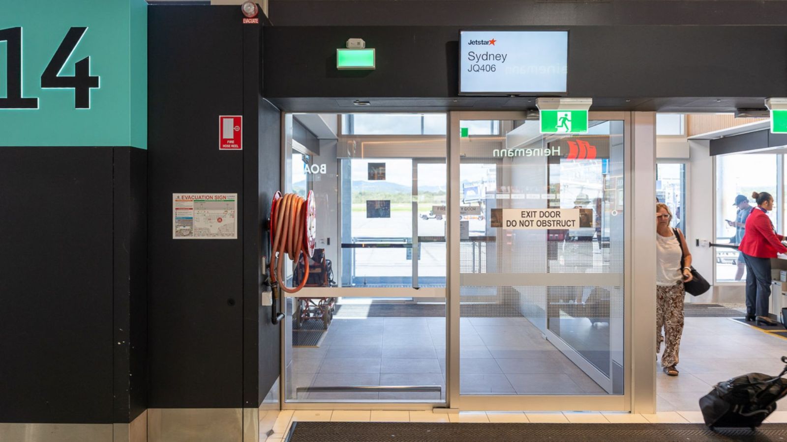 Airport boarding gate 14 with automatic glass doors leading to the Sydney flight. Signage above displays the destination and airline, while passengers enter the gate area. A fire hose reel and exit signs are mounted on the wall.