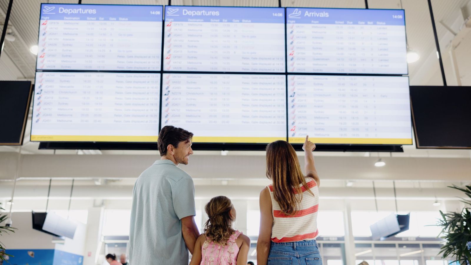 A family of three stands in front of a large digital flight information display at an airport. The mother points up at the departure and arrival screens while the father and daughter look on.
