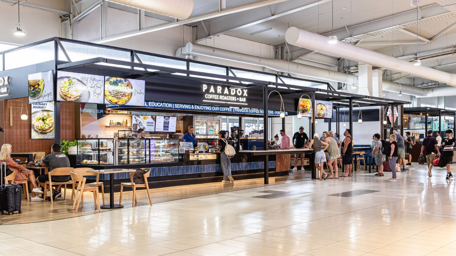 A busy airport dining area featuring the Paradox Coffee Roaster and Bar. The modern black-framed storefront displays digital menu boards with food images, while travellers line up or sit at tables enjoying their meals.