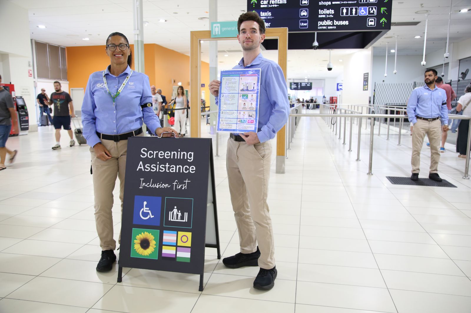 Two Gold Coast Airport staff members stand in a bright terminal near the security screening area, wearing blue shirts and beige trousers. One holds a sign that reads “Screening Assistance – Inclusion first” with accessibility and inclusion symbols, while the other holds an information poster. Queuing lanes and overhead wayfinding signs for taxis, public buses and toilets are visible in the background.