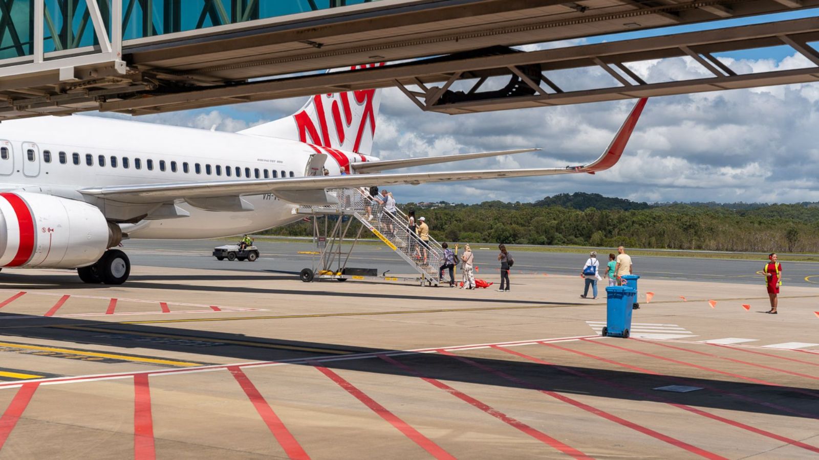 Passengers board a Virgin Australia aircraft using rear stairs on the tarmac at Gold Coast Airport. A jet bridge extends overhead, and ground staff assist with boarding as mountains and clouds fill the scenic background.