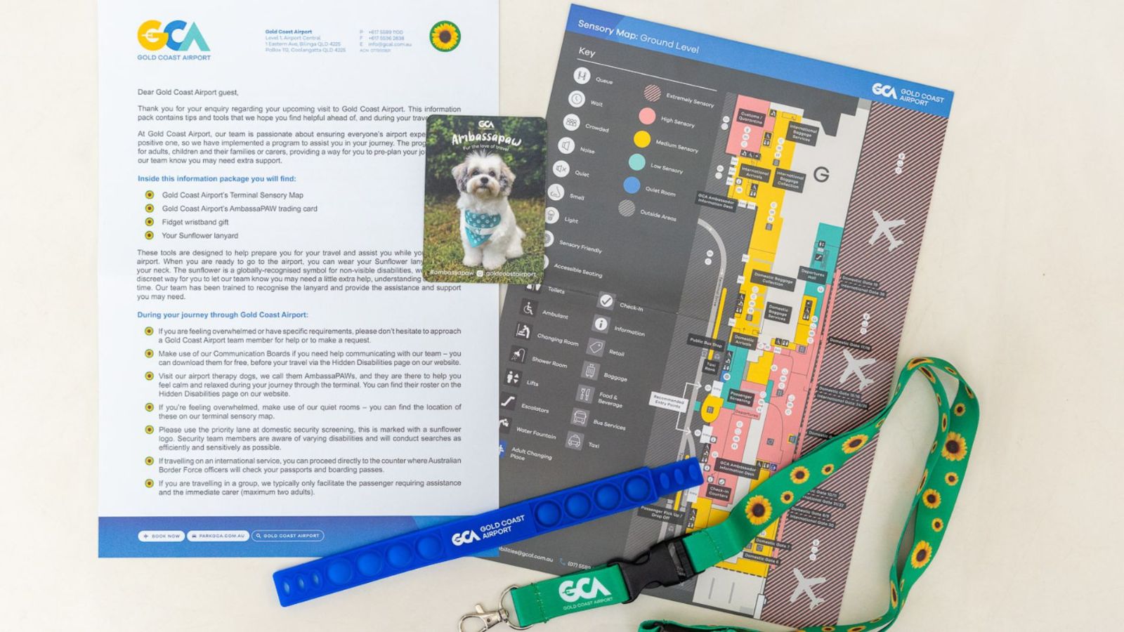 A Gold Coast Airport accessibility pack laid out on a surface, featuring an information sheet, terminal map, a photo card of a therapy dog, a Sunflower lanyard, and a blue pop fidget wristband marked “GCA Support.”