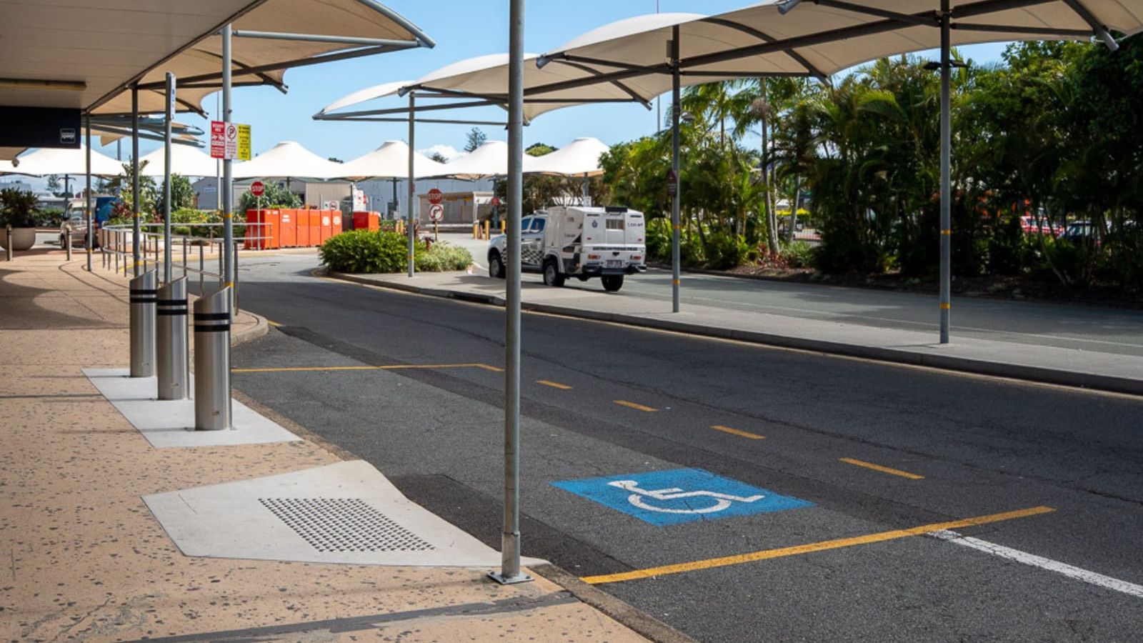 A designated accessible pick-up and drop-off zone at an airport, marked with a blue wheelchair symbol on the road. The area is covered by shade sails, with bollards lining the footpath and a vehicle driving in the background.