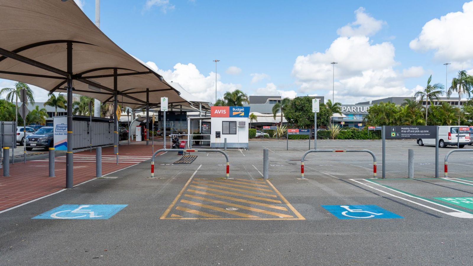Accessible parking bays at an airport car park, clearly marked with blue wheelchair symbols and space for rear access. Shade structures cover part of the area, with parking signs and a pay station visible in the background.