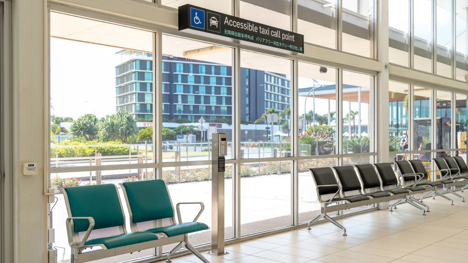 Accessible exit point inside an airport terminal, marked by a sign reading “Access to the Kerb Zone.” Green and black seating is arranged nearby, with large windows offering a view of nearby buildings and landscaped gardens outside.