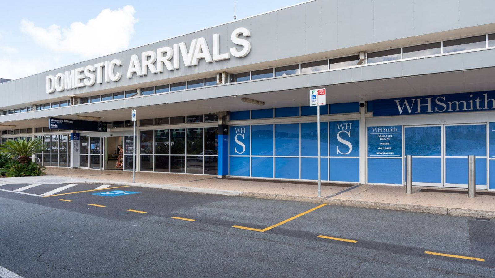 Exterior view of the Domestic Arrivals area at Gold Coast Airport, featuring clear signage above the entrance, a WHSmith storefront, and marked drop-off zones along the roadway.