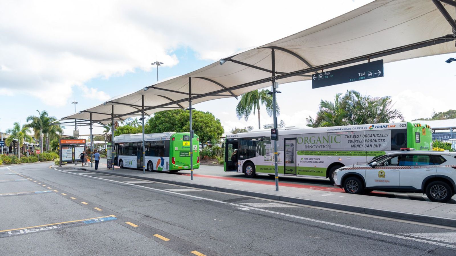 Airport bus and shuttle zone with multiple buses parked under large shade structures. A Gold Coast Airport vehicle is nearby and signs direct passengers to various transport services. Palm trees and blue skies complete the outdoor setting.