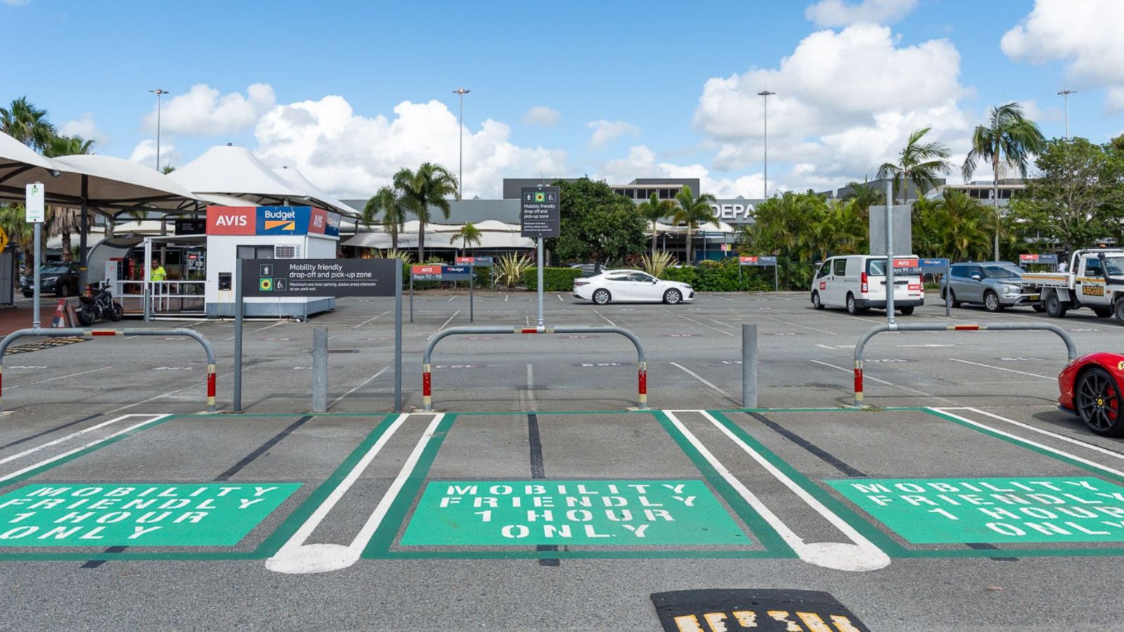 Dedicated mobility parking bays at an airport car park, marked with green paint and white text reading “Mobility Permit Holders Only.” The area includes bollards, signage, and nearby shade structures, with palm trees and vehicles in the background.