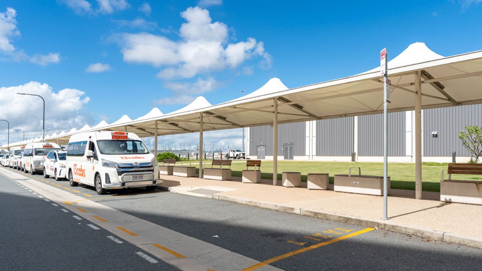 A row of white taxis parked at an airport taxi rank under large white shade structures. The area features benches, a footpath, and clear road markings, with a bright blue sky and scattered clouds overhead.