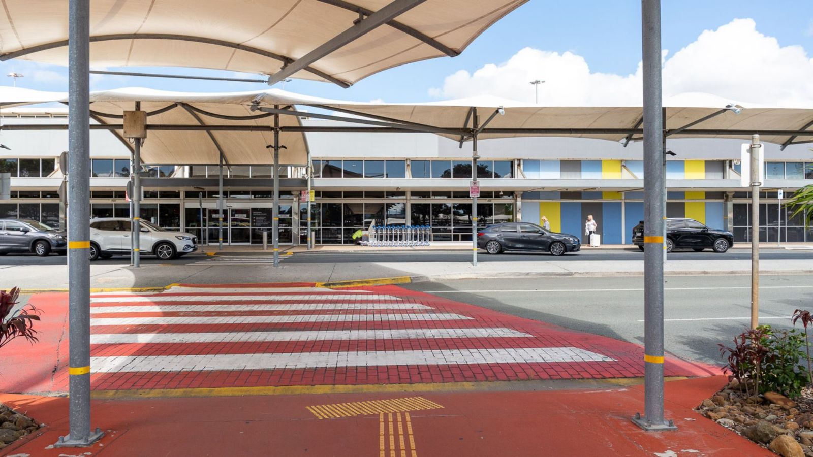 A red and white pedestrian crossing under shade sails at an airport, leading toward the terminal entrance. Yellow tactile paving and directional arrows enhance accessibility, with cars and buildings visible in the background.