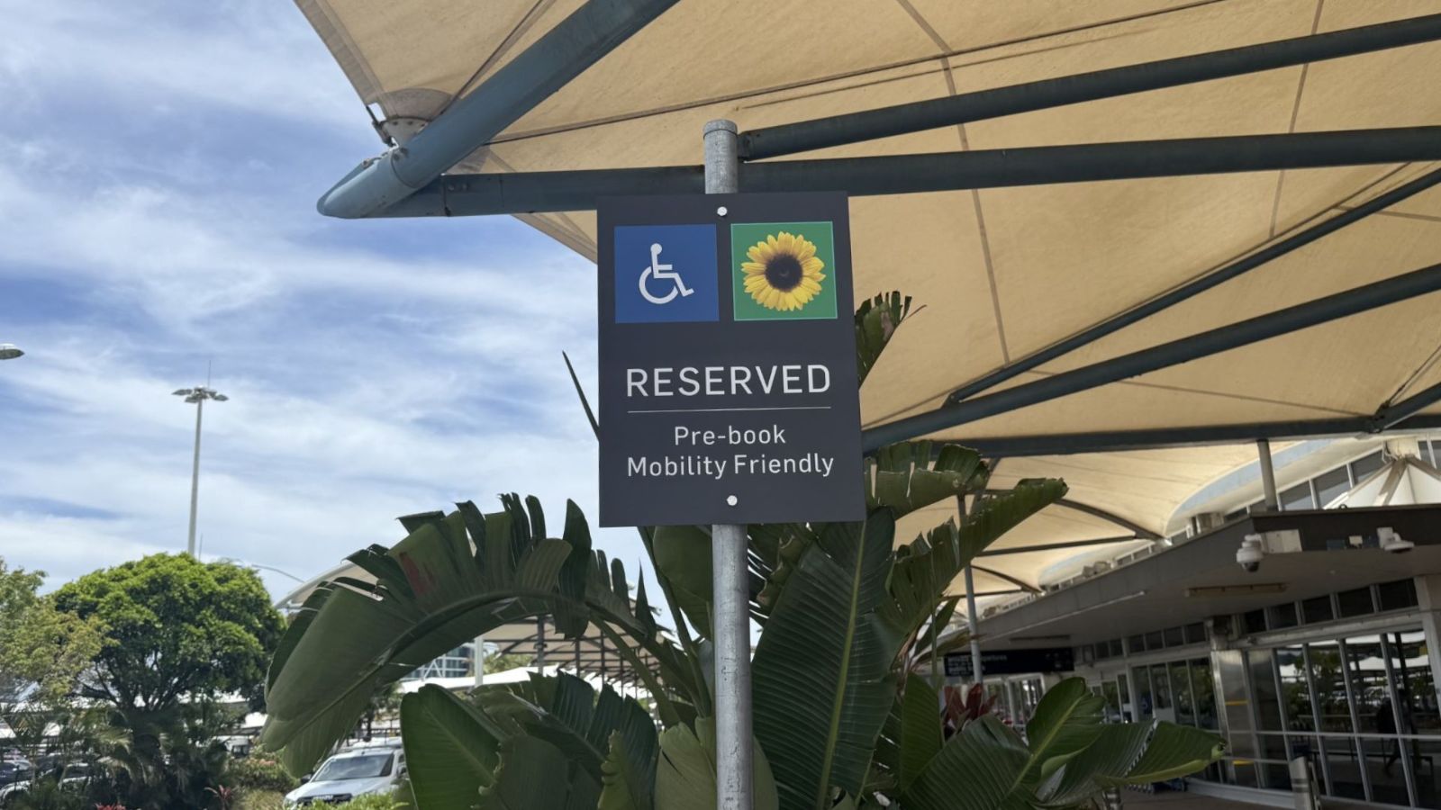 Reserved mobility-friendly parking sign with wheelchair and hidden disability sunflower symbol under a covered airport drop-off area.