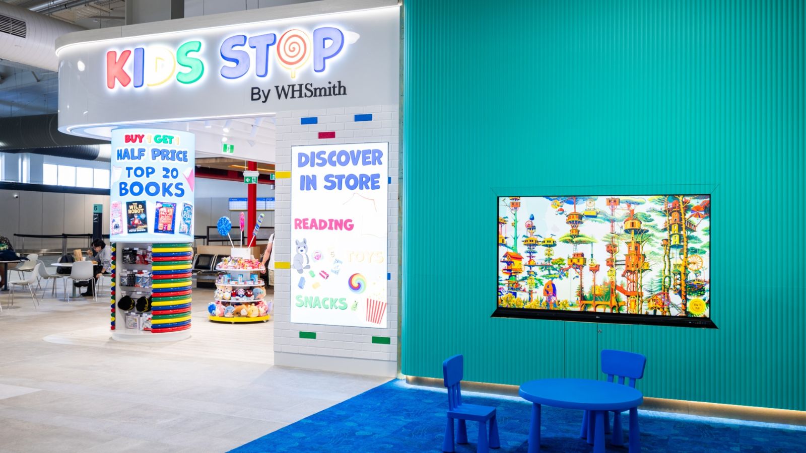 The colourful Kids Stop by WHSmith store at Gold Coast Airport, featuring a reading and play area with a small blue table and chairs, a bright wall-mounted screen, and signage promoting books and snacks.