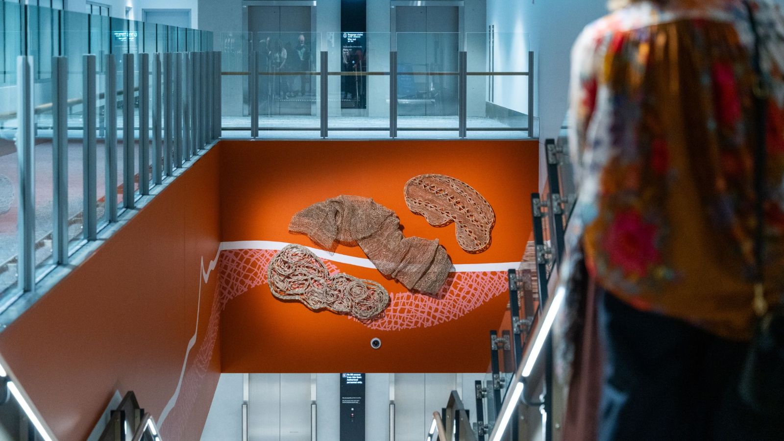 An installation in Gold Coast Airport’s International Arrivals Hall featuring woven sculptures made from native fibres by Casino Wake Up Time, a group of Bundjalung and Kamilaroi women. The artworks, set against a burnt orange backdrop with white patterns, symbolise the waterways of Cobaki Lakes and the surrounding land.