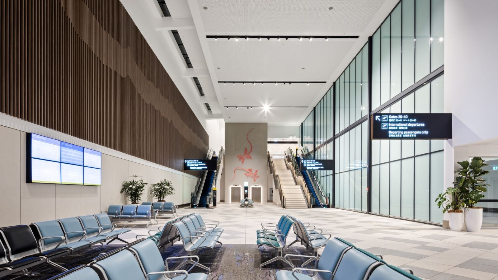 A spacious and modern airport terminal at Gold Coast Airport, featuring empty seating areas, digital flight screens, escalators, and red gecko wall art at the far end of the hall.