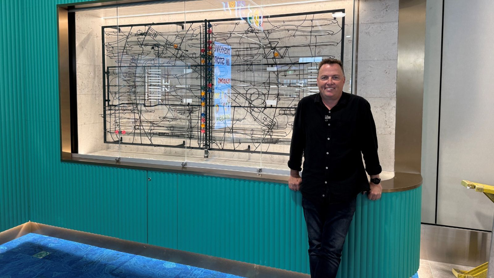A man stands smiling beside an intricate interactive wall installation at Gold Coast Airport, set against a teal feature wall near the Kids Stop by WHSmith area.