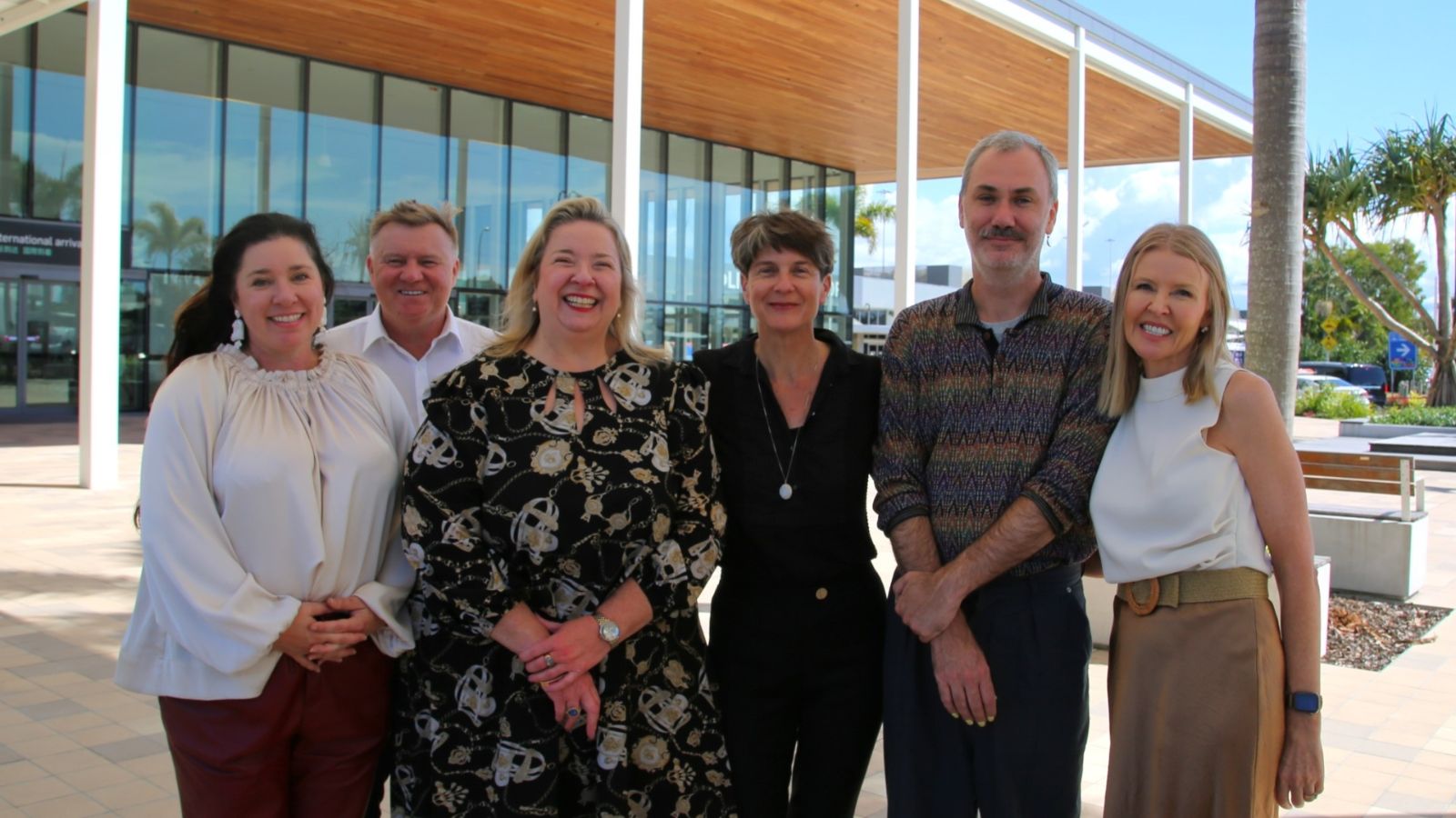 A group of six people stand together smiling outside Gold Coast Airport, with the terminal building and palm trees in the background on a sunny day.