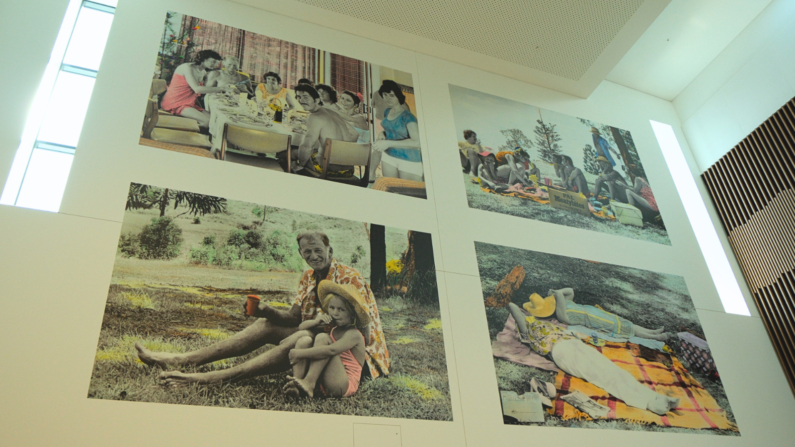 Large wall-mounted photographic artworks inside a modern building, featuring vintage-style family picnic and outdoor scenes with selective colour details.