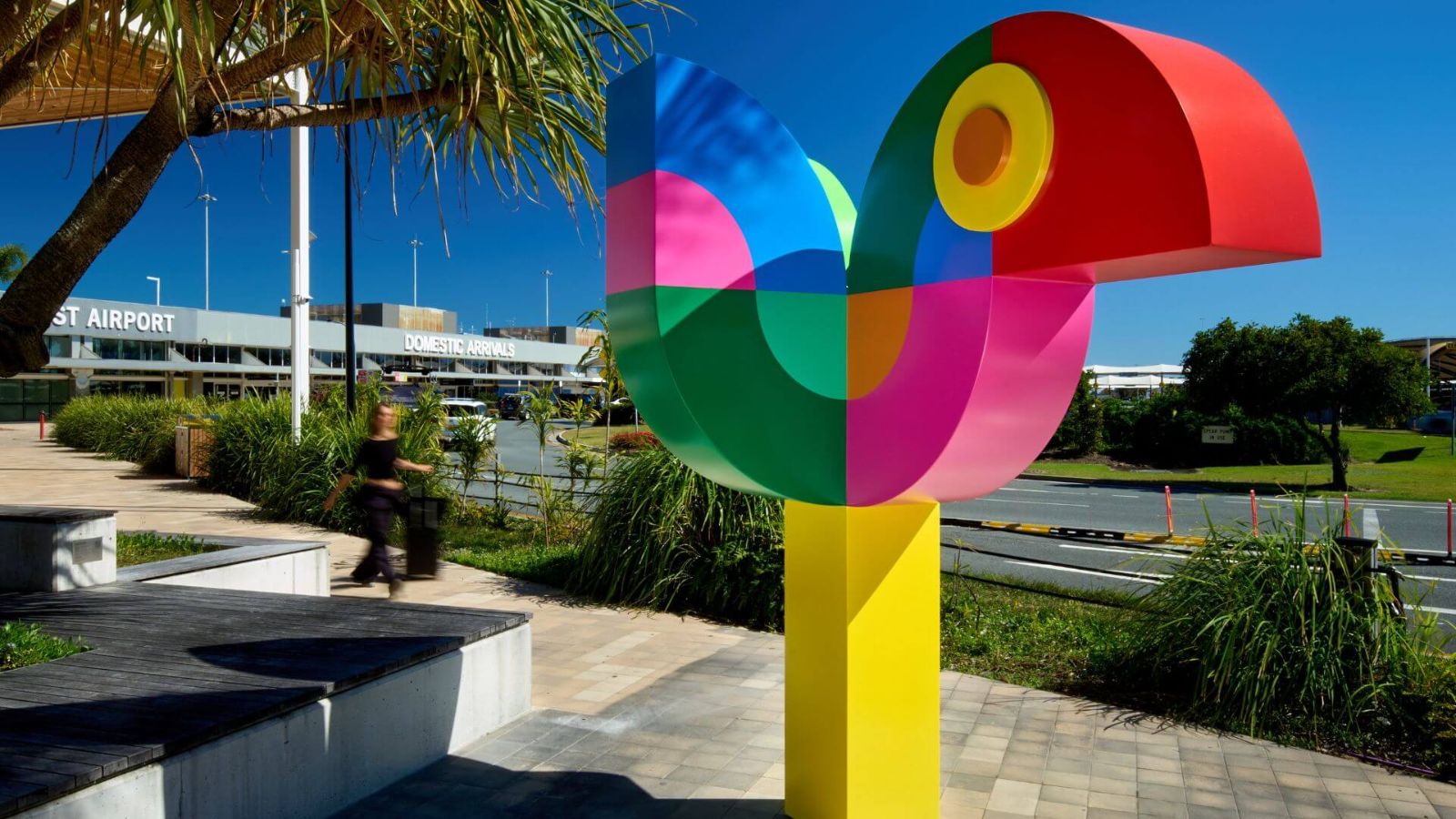 Brightly coloured abstract bird sculpture titled "Welcome Call" by Craig &amp; Karl, located outside Gold Coast Airport near the Domestic Arrivals entrance, with a clear blue sky in the background.