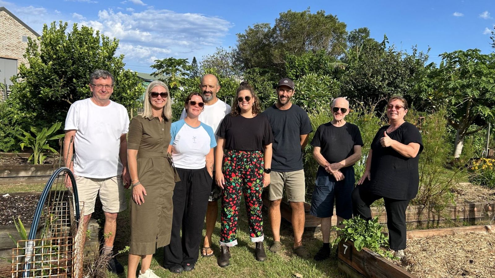 A group of eight people stands together in a lush community garden, smiling at the camera. The setting includes raised garden beds, vibrant greenery, and a clear blue sky. The group is diverse in age and attire, with some wearing casual clothing and sunglasses. One person on the right gives a thumbs-up, adding a welcoming and positive feel. The background features trees, plants, and a brick building, indicating a suburban or urban gardening space.