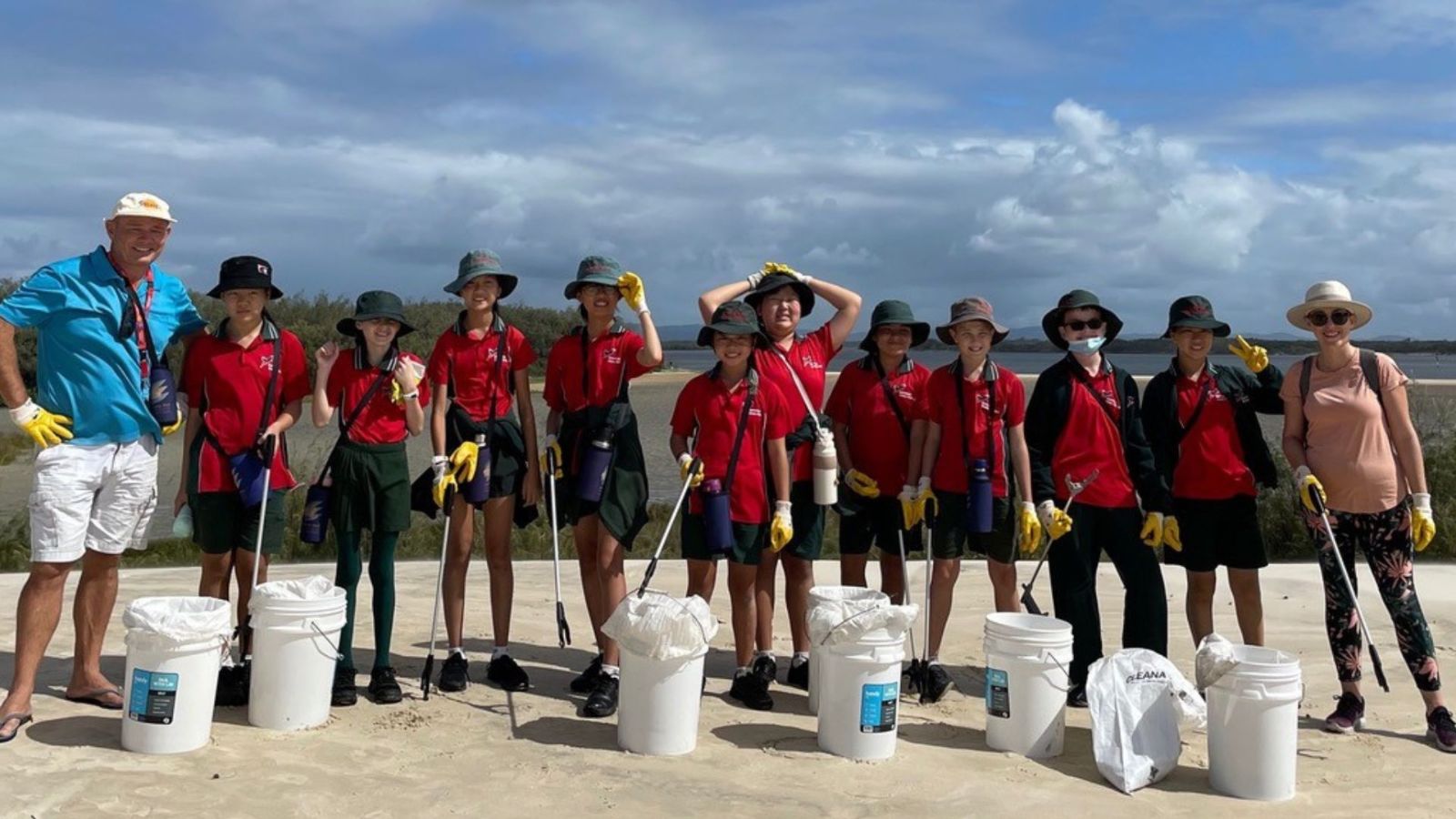 A group of students in red and green uniforms, along with two adult supervisors, participate in a beach cleanup as part of a marine conservation project. They are standing on sandy terrain near a coastal area, holding litter pickers and buckets while wearing wide-brimmed hats, gloves, and hydration packs. The sky is partly cloudy, and the background features coastal vegetation and a tidal inlet. The group appears engaged and enthusiastic about their environmental efforts.