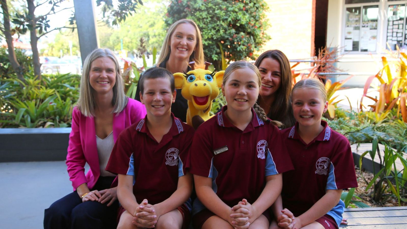A group of three smiling school students in maroon and blue uniforms sits in the front row, with three women in professional attire sitting behind them. A bright yellow giraffe mascot, Healthy Harold, is positioned in the middle, adding a fun and engaging element. The photo is taken outdoors in a school courtyard with lush greenery, a modern building, and a bulletin board visible in the background. The group appears happy and engaged, likely part of an educational event promoting health and well-being.
