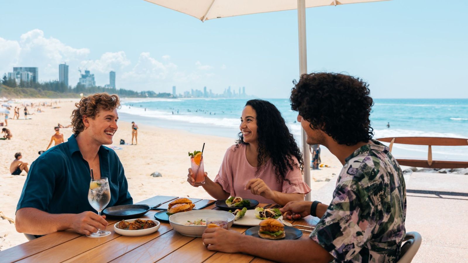 Outdoor dining at Rick Shores in Burleigh Heads with a wooden table set with gourmet dishes and drinks, overlooking the sandy beach and turquoise ocean with Surfers Paradise skyline in the distance under a clear blue sky.