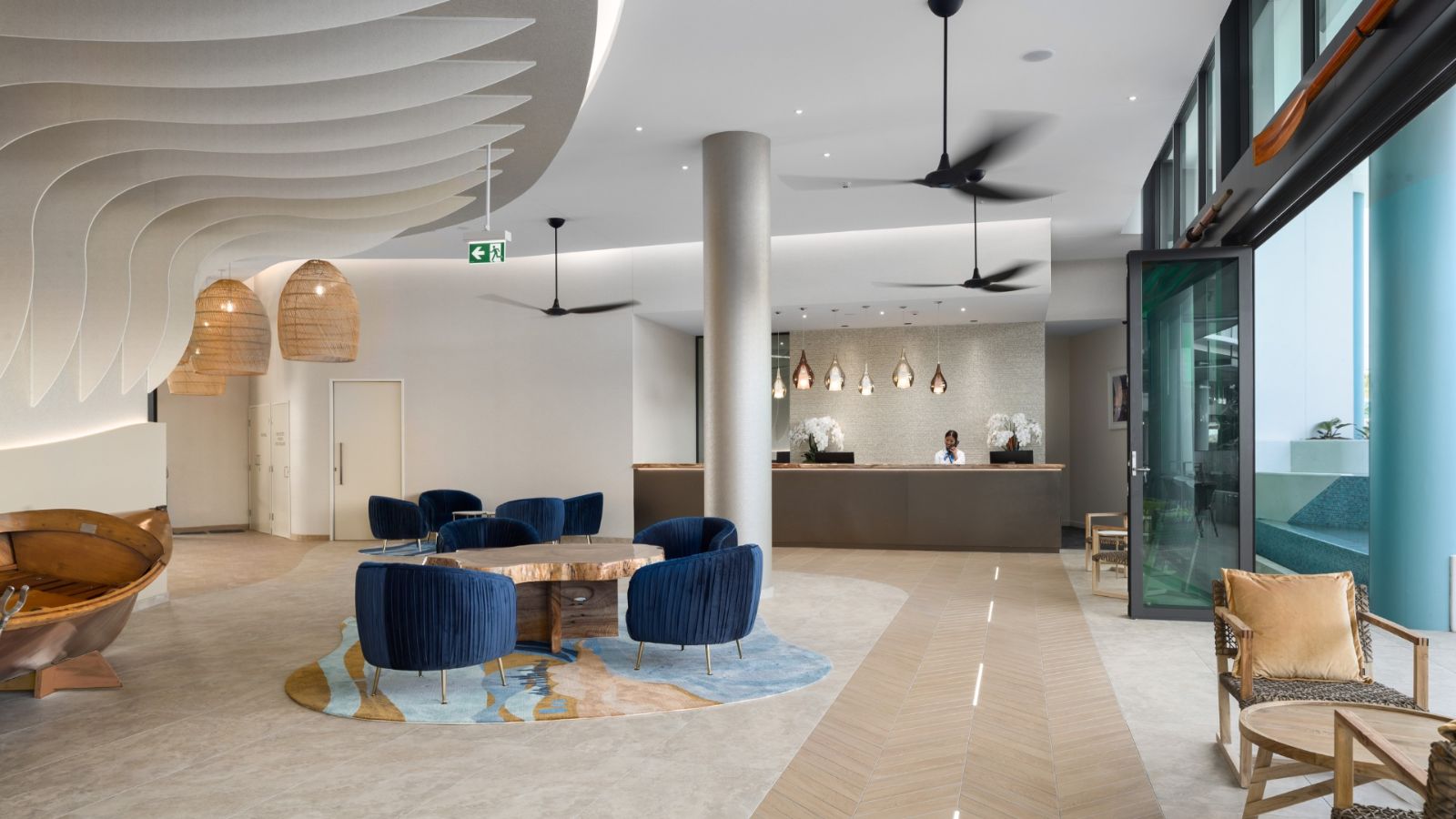 Modern lobby of Rydges Gold Coast Airport featuring a stylish curved ceiling design, blue velvet chairs around a wooden table on a patterned rug, natural timber accents, and a reception desk with pendant lighting.