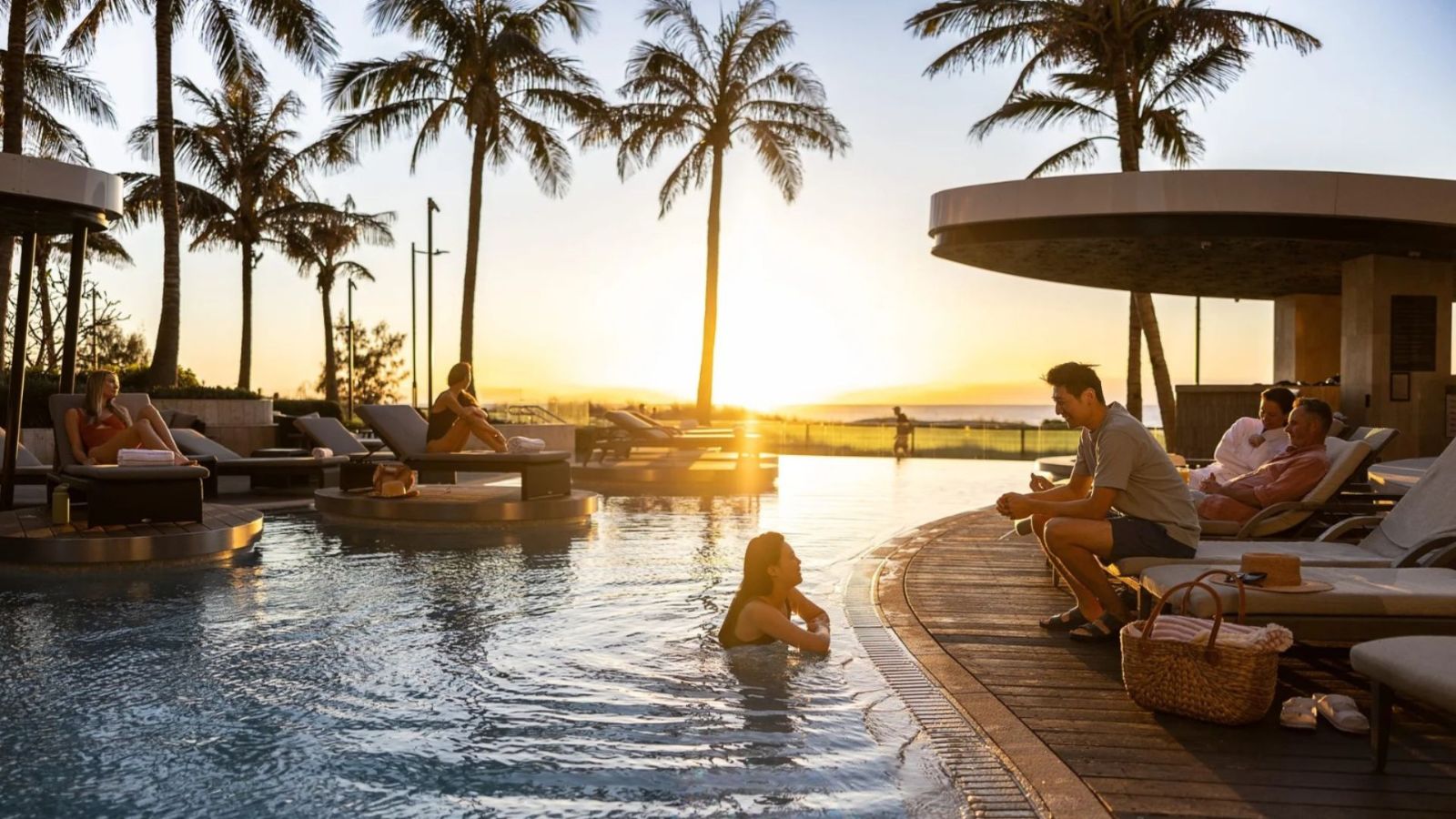 Luxury poolside setting at The Langham Hotel, Gold Coast during sunset, featuring palm trees, sun loungers, and guests relaxing by the water with ocean views in the background.<br/>