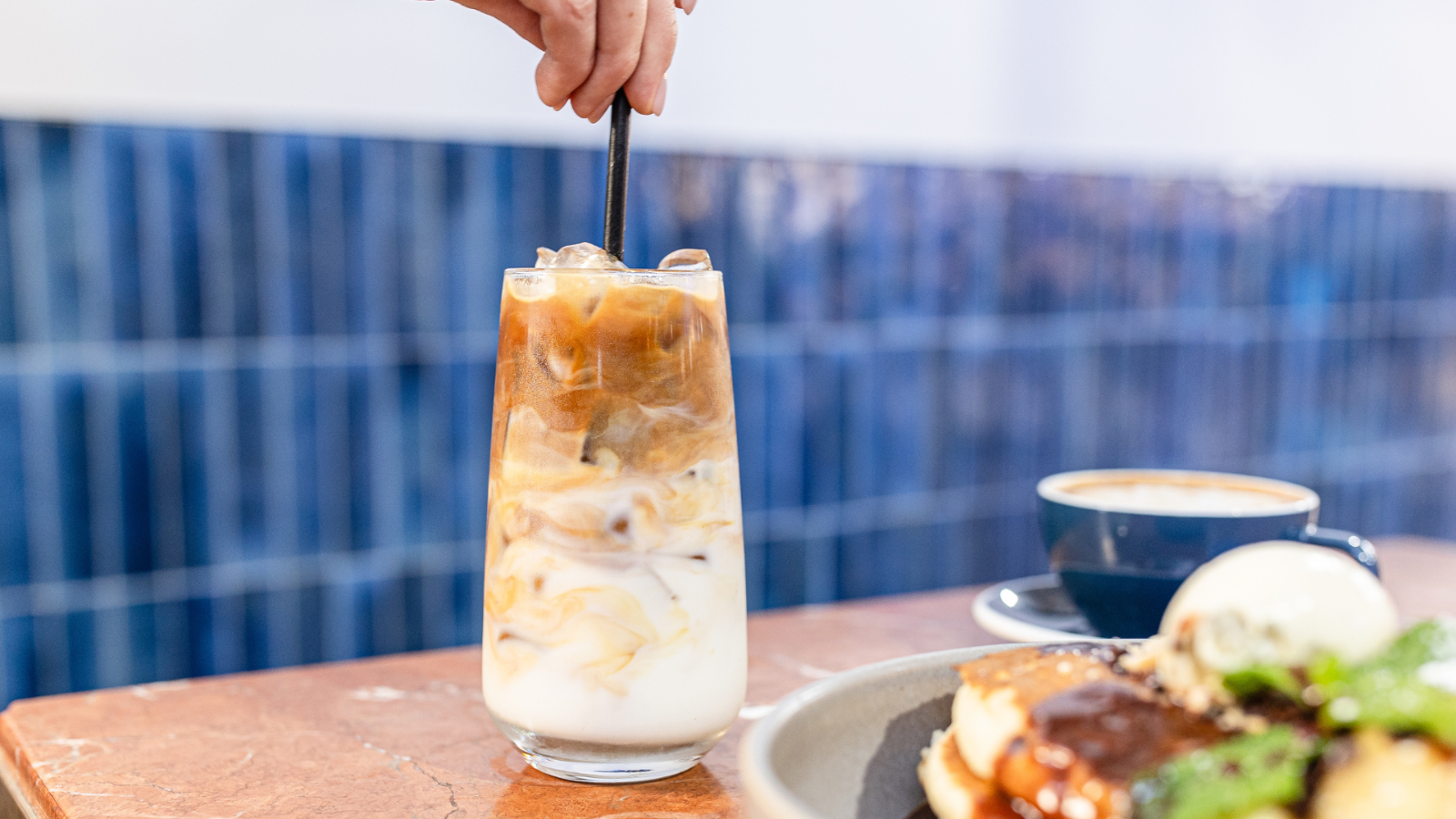 Hand stirring iced latte in a tall glass with milk and coffee layers on a café table, with a cappuccino and plated dessert blurred in the foreground.
