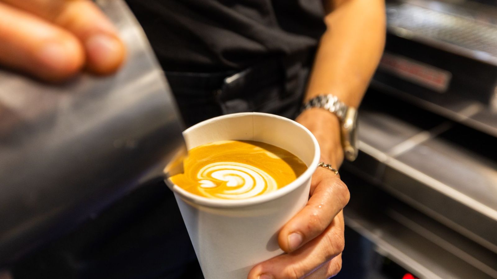 Barista pouring steamed milk into a takeaway cup of coffee to create latte art, with a swirl pattern forming on the surface.