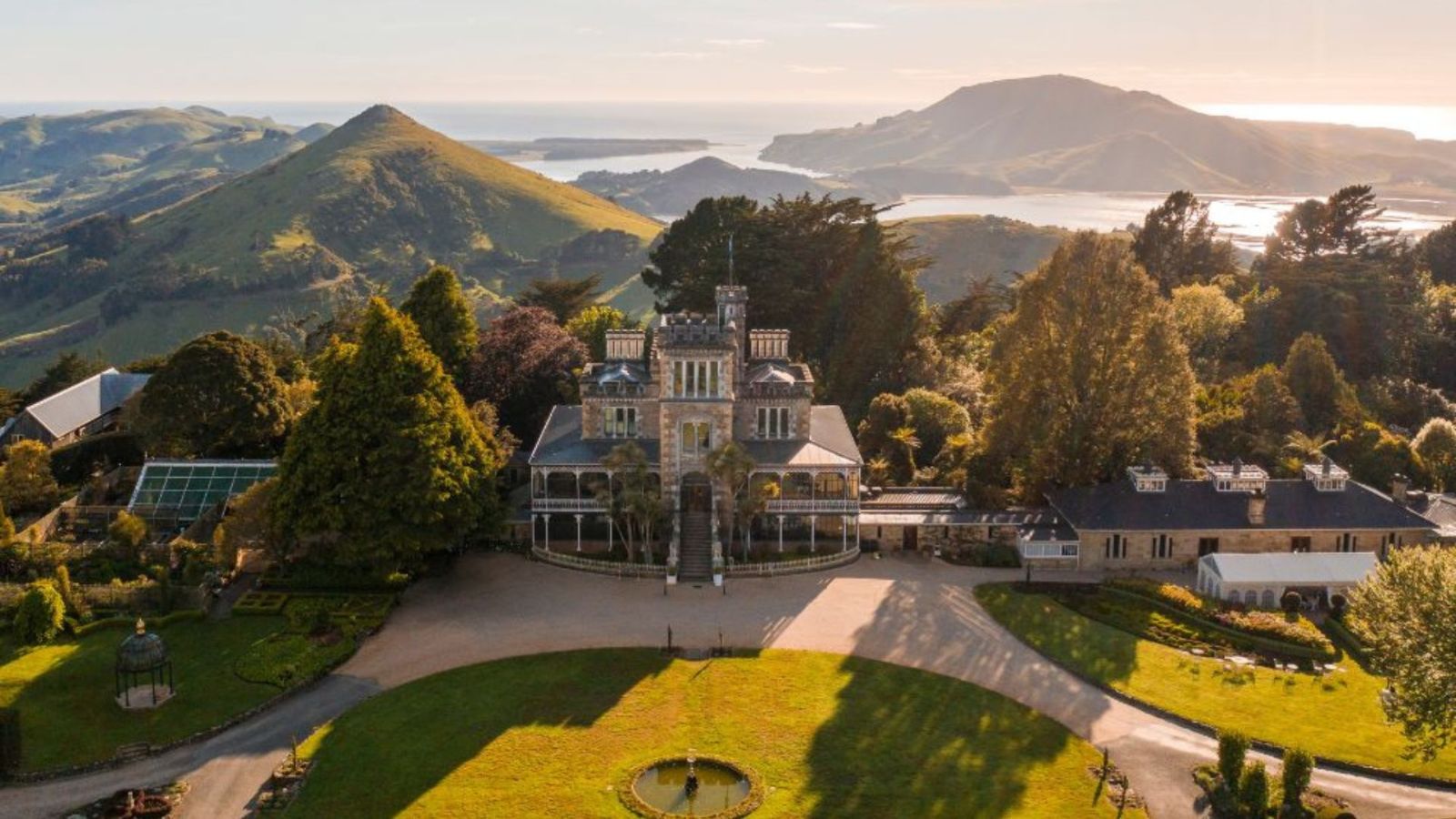 Aerial view of Larnach Castle surrounded by lush gardens on the Otago Peninsula, with rolling green hills and the Pacific Ocean in the background under a clear sky.
