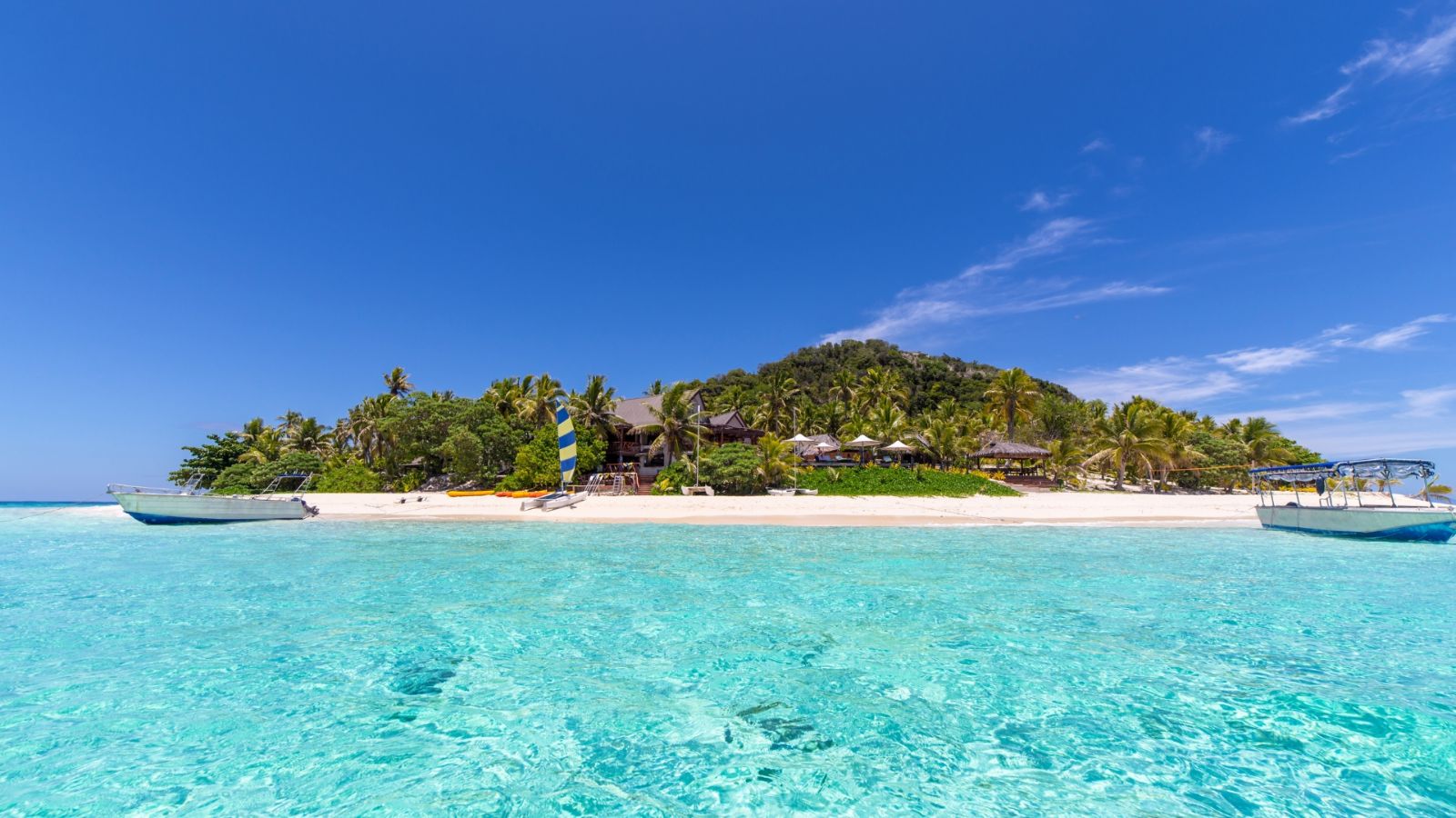 Tropical island beach in Fiji with clear turquoise water, white sand, palm trees and boats anchored offshore.