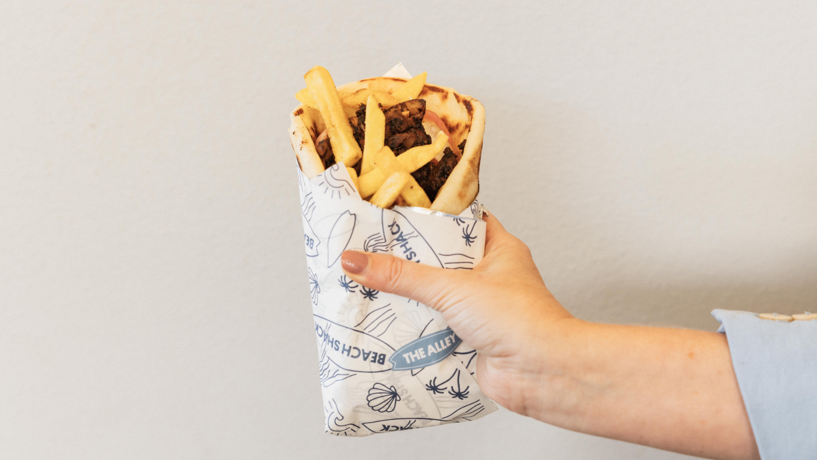 Hand holding a wrapped yiros filled with grilled meat and French fries in branded paper against a plain light-colored background.