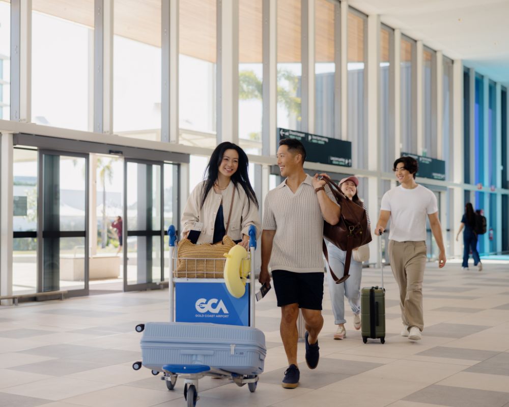 Passengers walking through the arrivals hall at Gold Coast Airport with luggage trolleys and suitcases, under natural light streaming through tall windows.